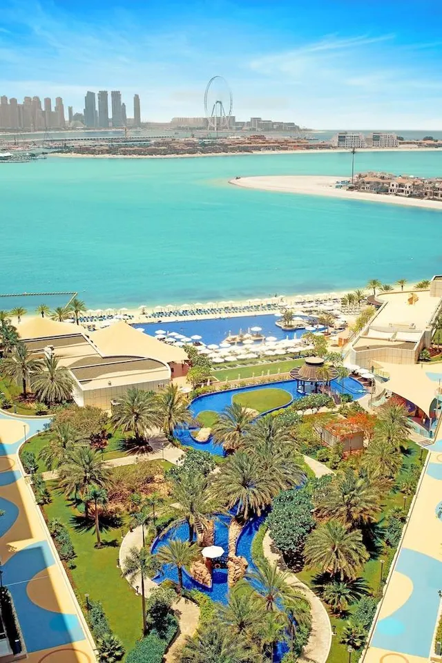 Aerial view of a resort with pools, palm trees and umbrellas beside a turquoise sea, with the Dubai skyline and the Ain Dubai ferris wheel in the distance.