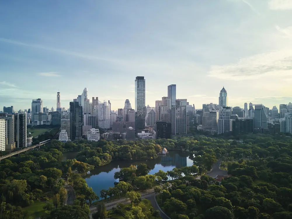 Elevated view of the Bangkok skyline beyond a green park with a lake and an elevated train line on the left.