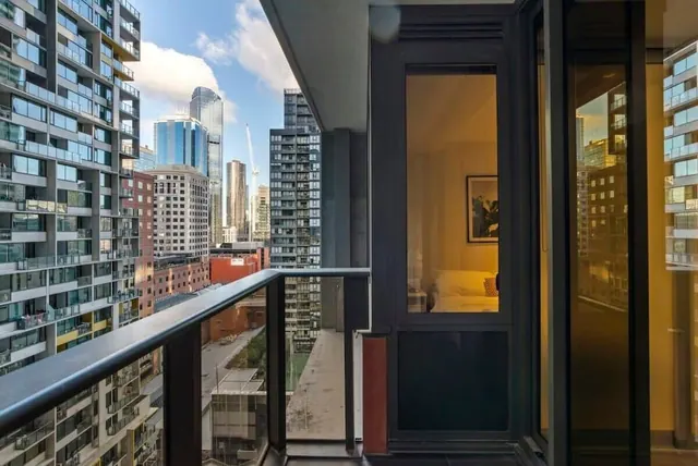 A glass-railed balcony looks out over high-rise towers and a city skyline, with a bedroom visible through the glass door.