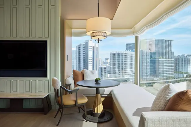 A seating corner with a banquette, round table and chairs beside a large window showing high-rise buildings under a blue sky, with a wall-mounted television to the left.