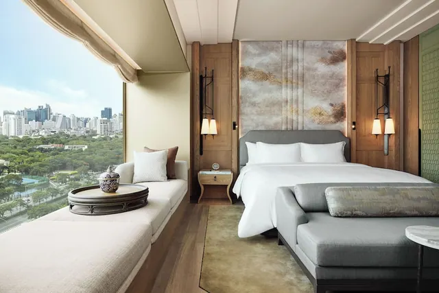 A bedroom with a king bed, a grey bench at the foot of the bed and a window seat, with a city skyline visible through the large window.