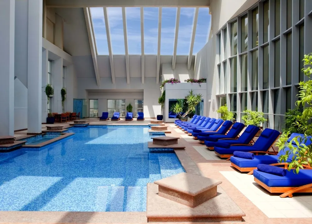 An indoor pool with blue-cushioned sun loungers along one side, under a skylit ceiling with tall windows on the right.