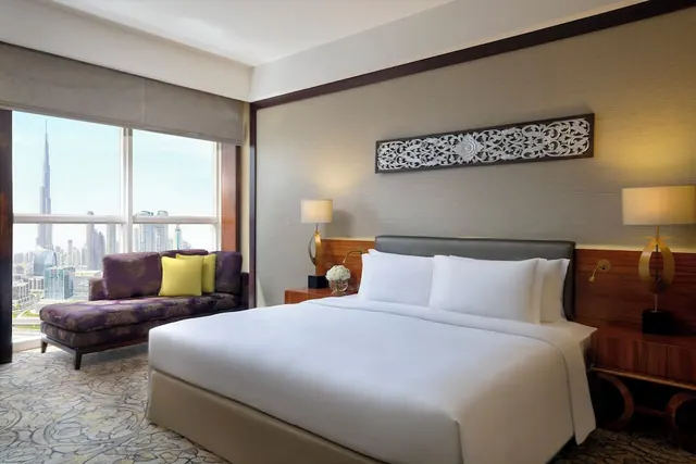 A made-up king bed faces a purple sofa and a large window with a view of the Burj Khalifa tower and city buildings.