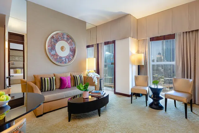 A hotel suite living area with a sofa, armchairs, a coffee table and large windows overlooking high-rise towers.