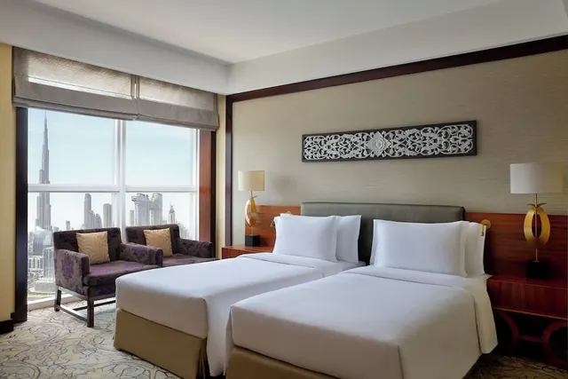 A bedroom with two white twin beds and two armchairs by a window, with the Burj Khalifa visible in the skyline outside.