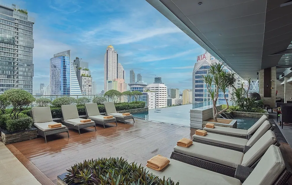A rooftop pool deck with cushioned sun loungers and folded towels beside an infinity-edge pool, with city high-rises visible under a partly cloudy sky.