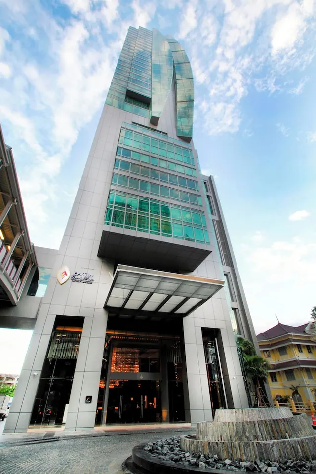 Low-angle view of a tall glass-clad hotel tower with a covered entrance canopy, a stone planter in front and a yellow building to the right under a partly cloudy sky.