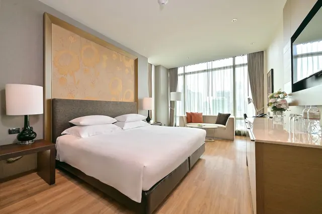 A hotel bedroom with a king bed, a seating area by floor-to-ceiling windows, and a wall-mounted television.