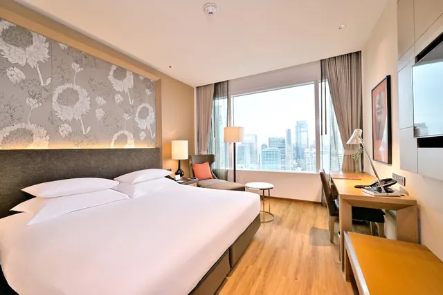 A hotel bedroom with a king bed, armchair, floor lamp, work desk and a large window overlooking city high-rise towers.