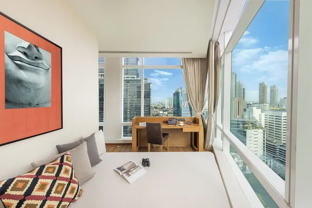 A bright room with a long white daybed, a wooden desk and chair, and floor-to-ceiling windows overlooking city towers.