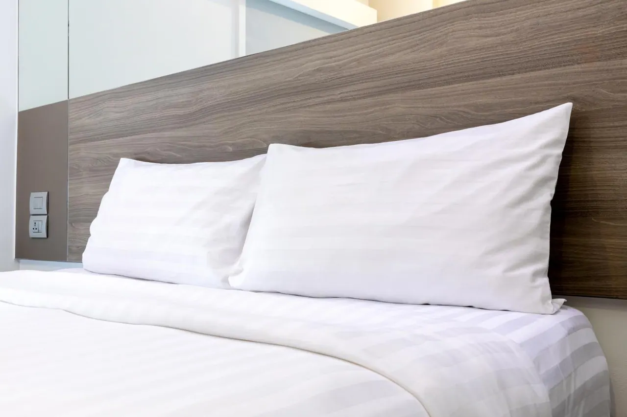 A made bed with white striped linen and two pillows sits in front of a wooden headboard, with a wall switch panel on the left.