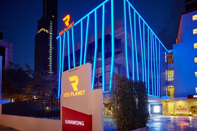 Night exterior of the Red Planet Surawong hotel with blue neon lines running up the facade and illuminated Red Planet signage at the entrance.