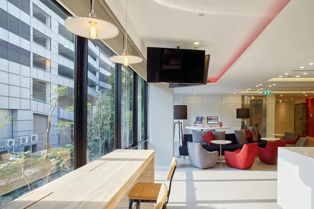 A lounge area with a long wooden table by floor-to-ceiling windows, red and grey armchairs, a wall-mounted TV, and a desk with computers, looking out onto a neighbouring building and trees.