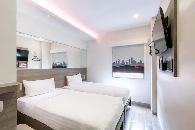 Twin beds with white linen in a bedroom with a wall-mounted television, a mirrored wall, and a printed skyline roller blind over the window.