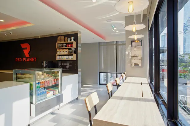 A café counter with a glass drinks fridge and coffee machine sits beside a long wooden communal table and chairs near floor-to-ceiling windows overlooking city buildings.