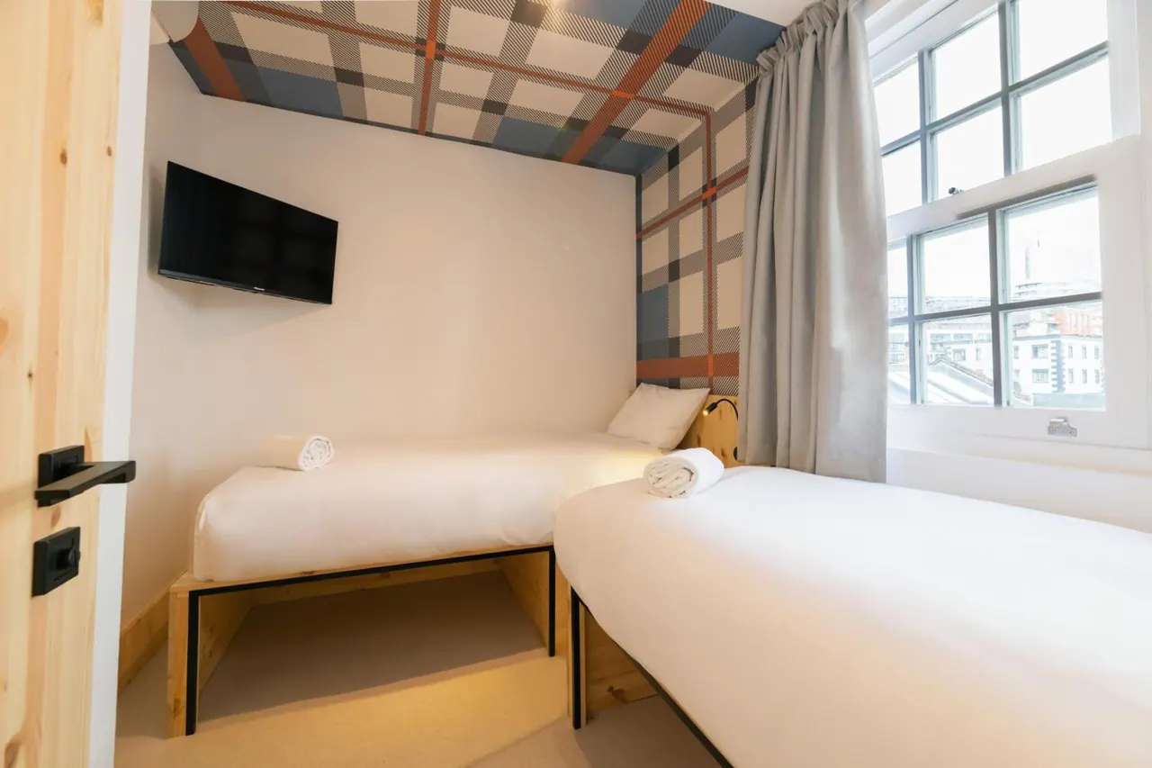 A small bedroom with two single beds, a wall-mounted television, a tartan-patterned ceiling and a window looking out over city rooftops.