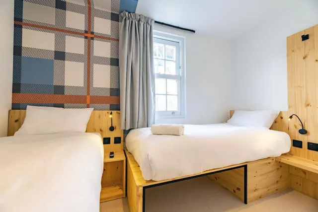 A bedroom with two single beds with white bedding, a tartan-patterned feature wall, a sash window and pine wood headboards.
