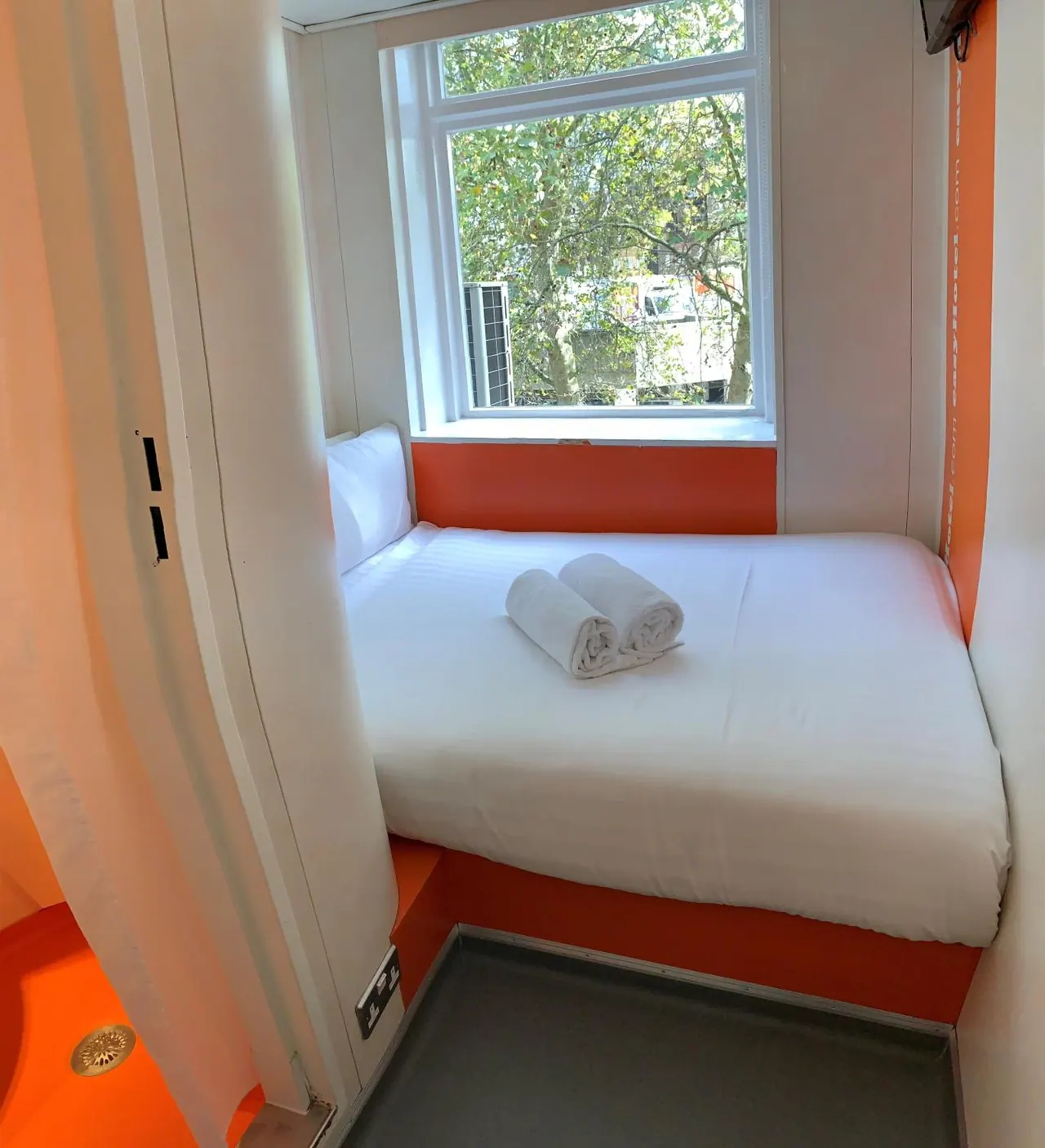 A small bedroom with a made double bed, two rolled white towels on the duvet, orange walls, and a window showing trees outside.