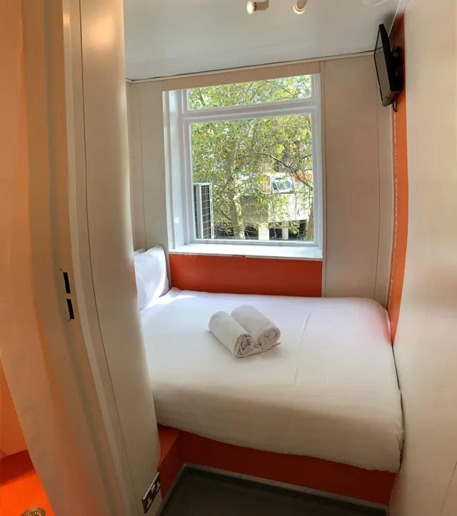 A small bedroom with a double bed, two rolled white towels on the bed, a window looking out onto trees, and a wall-mounted television.