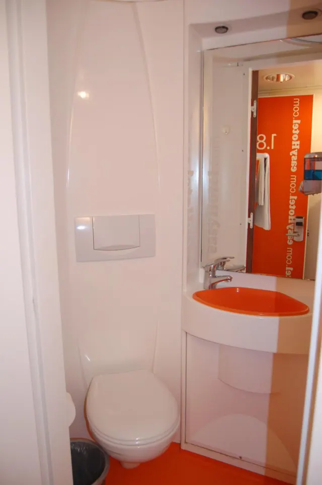 A small bathroom with a wall-mounted toilet, an orange basin beneath a mirror that reflects an orange wall with easyHotel branding, and a bin on the floor.