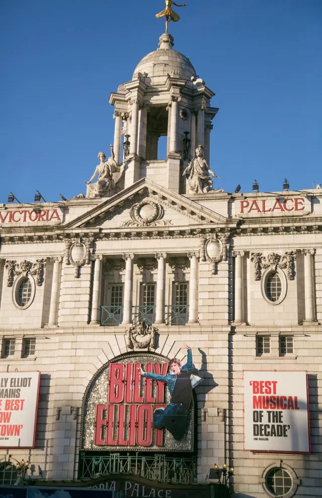 The stone facade of the Victoria Palace Theatre, with a domed tower topped by a gilded statue, a Billy Elliot banner and a Best Musical poster, under a clear blue sky.