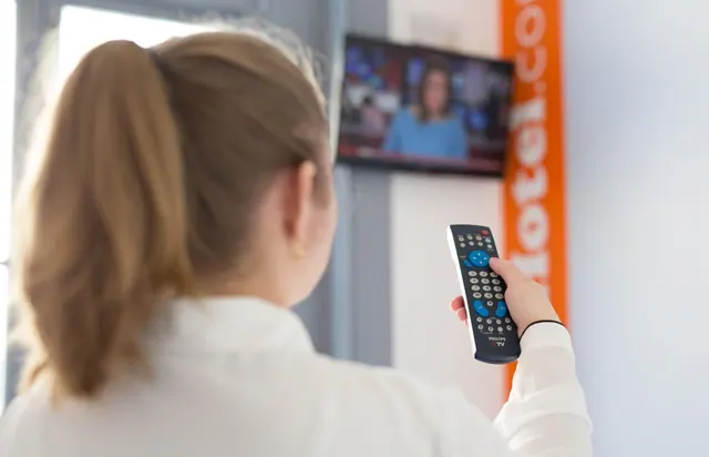 A woman with her back to the camera points a Philips remote at a wall-mounted television showing a news presenter, beside an orange banner with partly visible hotel lettering.