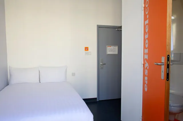 A small bedroom with a made double bed and white pillows, beside a grey door and an open orange door that shows a toilet in the bathroom beyond.