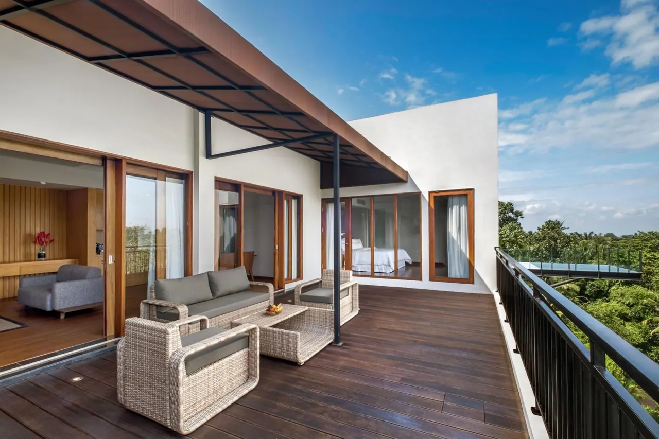 A covered wooden deck terrace with wicker seating opens through glass doors to a room with a bed, and trees and a pool are visible beyond the railing.