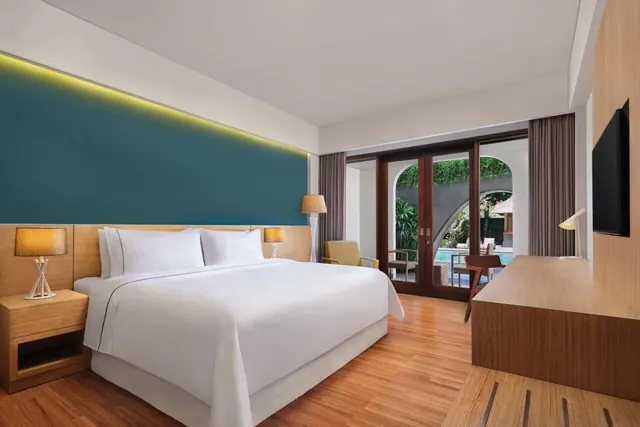 A bedroom with a made king bed, a teal accent wall, a wooden desk and glass doors opening onto a pool and greenery.