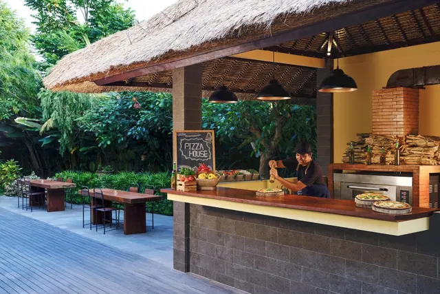 An outdoor pizza counter with a thatched roof, a brick oven and a chef at work, with wooden dining tables on the deck beside it.