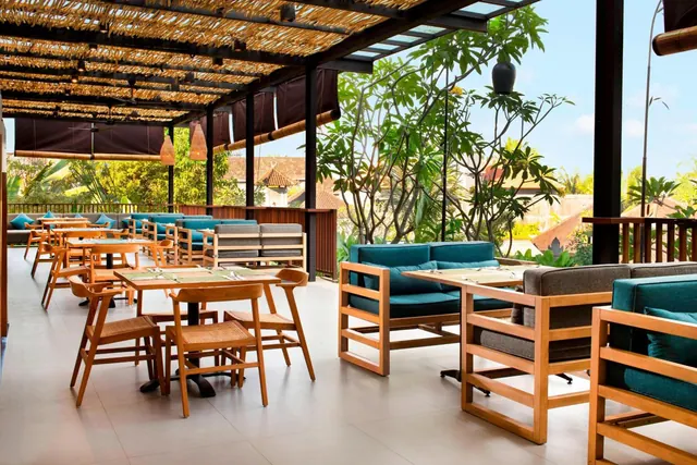 An outdoor restaurant terrace with wooden tables and chairs and teal sofas under a thatched pergola, with trees and rooftops visible beyond the railing.