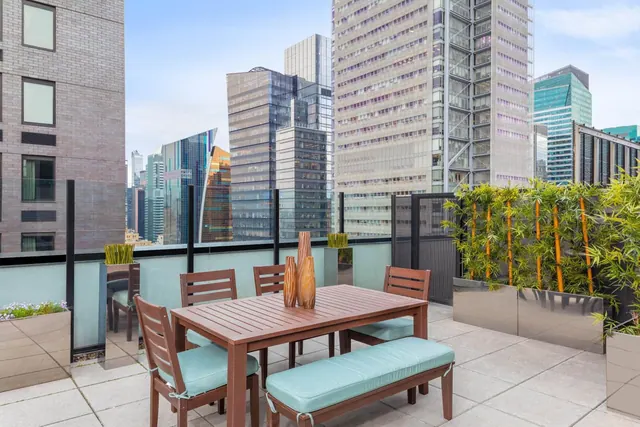A furnished rooftop terrace with a wooden dining table, chairs and bench with teal cushions, bamboo planters, and high-rise office buildings in view.