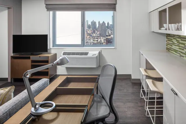 A hotel room with a desk and lamp, a television on a wooden cabinet, a kitchenette counter with bar stools, and a window showing a city skyline.