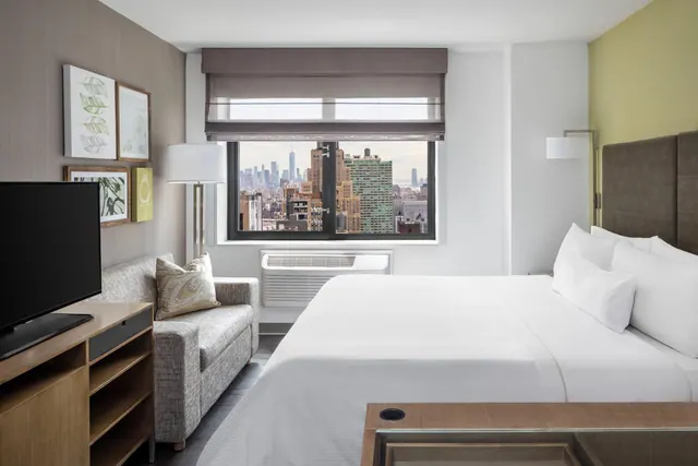 A bedroom with a king bed, a grey sofa, a wall-mounted television and a window looking out over a city skyline.