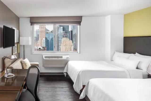 A hotel bedroom with two made beds, a wall-mounted television, a desk with a chair, and a window overlooking city skyscrapers.
