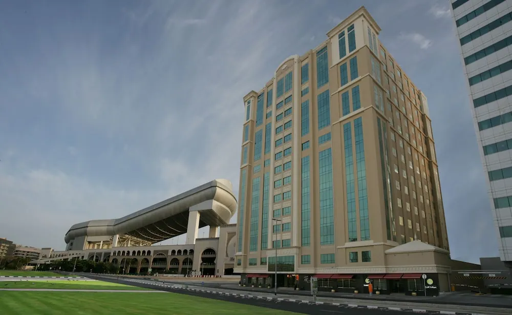 Exterior view of a tall beige hotel tower with teal glass windows, a curved arched structure beside it, and a green lawn and road in the foreground.