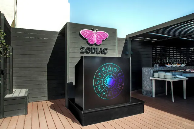 An outdoor terrace with a Zodiac sign and pink butterfly logo, a black cube displaying a zodiac wheel, and a serving table with stacked plates under a pergola.