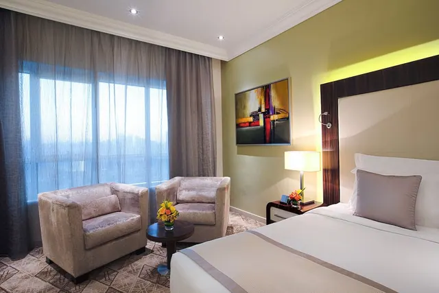 A hotel bedroom with a large bed, two armchairs beside a sheer-curtained window, a small round side table with flowers, and an abstract painting on the wall.