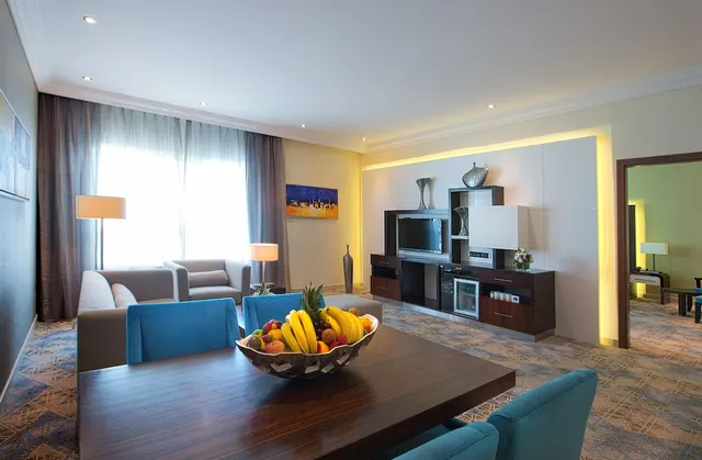 A hotel suite living room with grey sofas, blue armchairs, a fruit bowl on a dark wooden table, and a wall unit holding a television and small fridge.