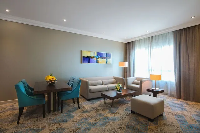 A furnished living area with a sofa, armchair, coffee table, a dining table with a fruit bowl and teal chairs, and a large window with sheer curtains.