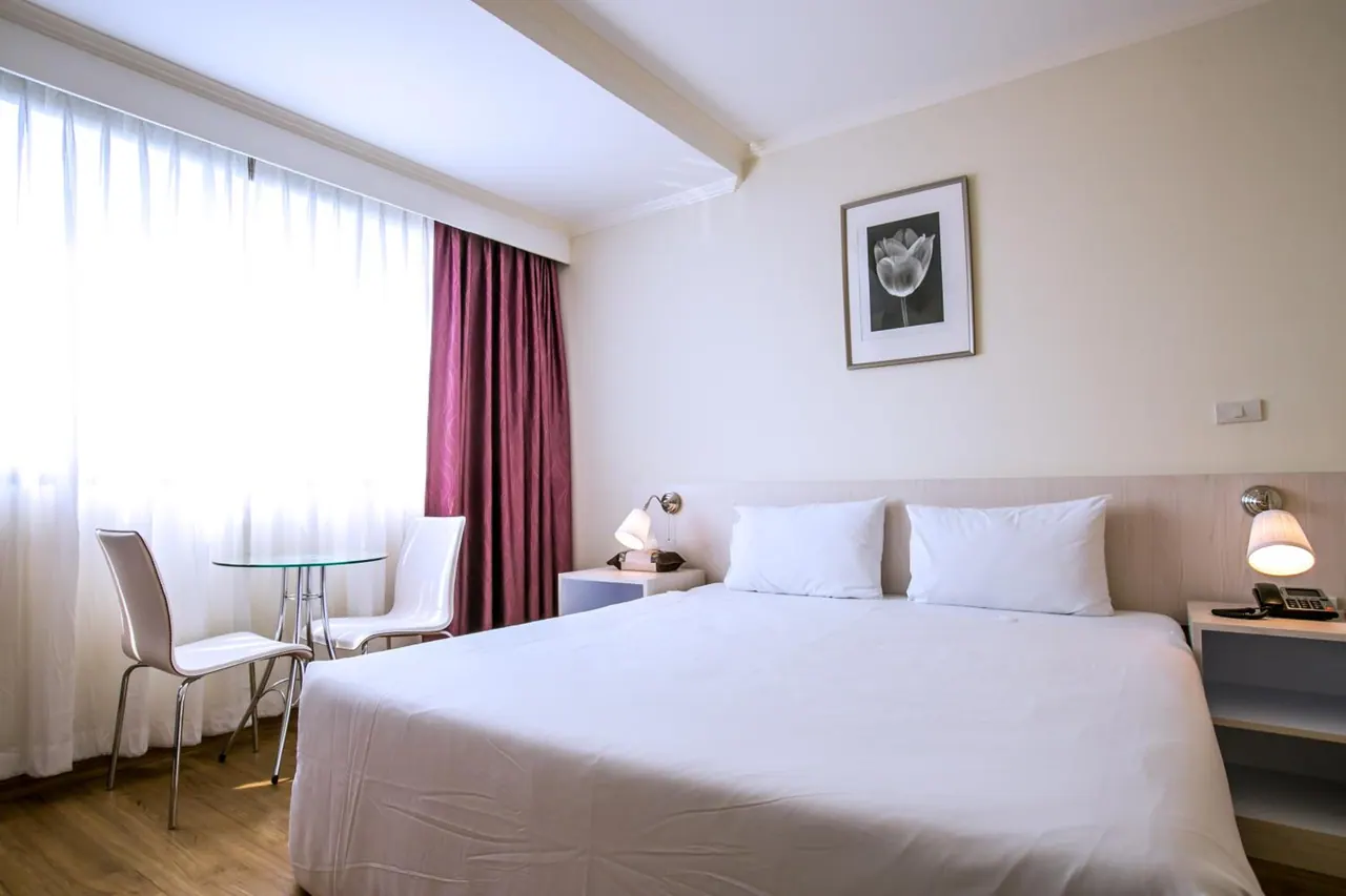 A bedroom with a king bed, bedside lamps and a telephone, and a small glass table with two white chairs beside a sheer-curtained window.