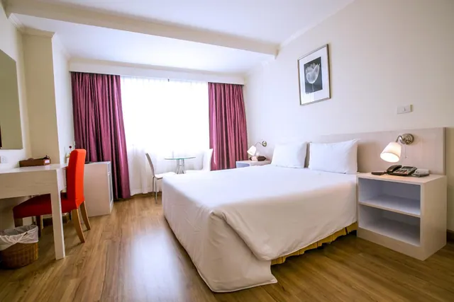 A hotel bedroom with a double bed under white bedding, a desk with a red chair, a small round table by the curtained window, and bedside lamps.
