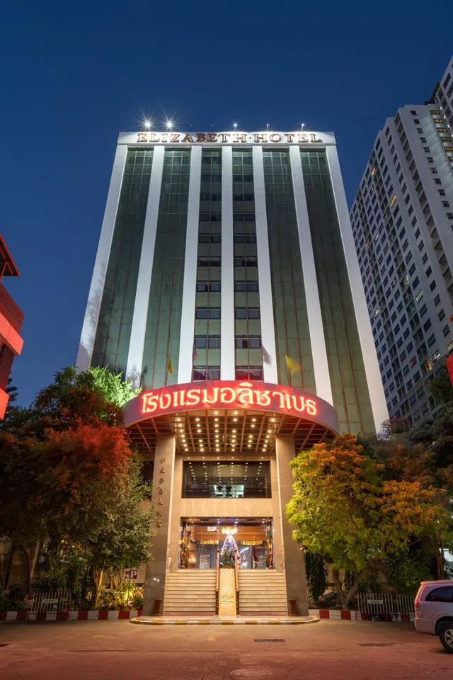 The Elizabeth Hotel tower at dusk, with its lit rooftop sign, red Thai-script entrance arch, and trees along the street.