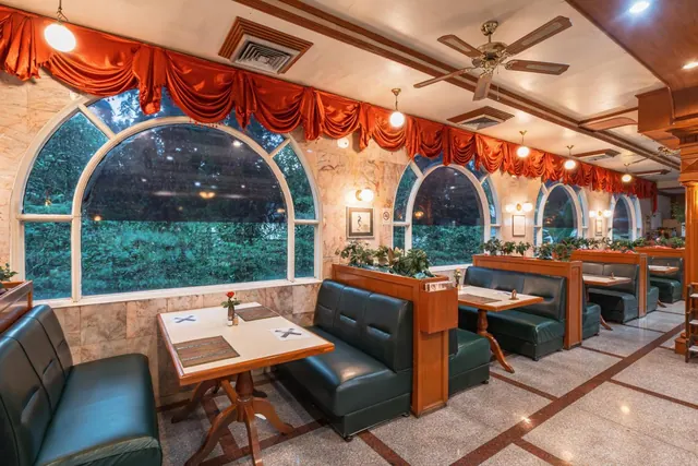 A dining room with green leather booths, wooden tables, and arched windows looking out onto trees and greenery, with red drapes along the top of the windows.