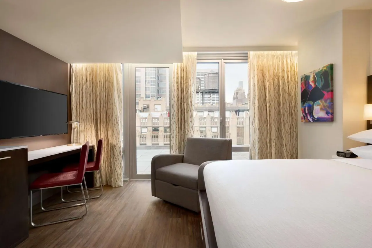 A hotel bedroom with a large bed, a desk with red chairs, a wall-mounted television, an armchair, and glass doors opening onto a terrace with city buildings beyond.