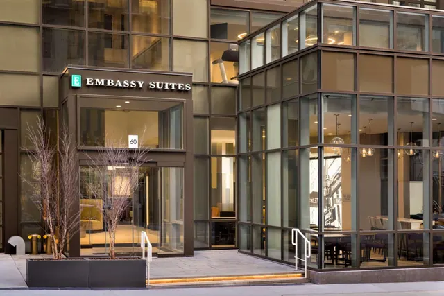 The entrance of an Embassy Suites hotel shows a glass facade, an illuminated sign reading EMBASSY SUITES, the number 60, planters with bare trees, and steps with metal handrails.