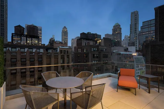 A rooftop terrace at dusk has a round table with four mesh chairs, an orange lounge chair and a side table, overlooking high-rise buildings.