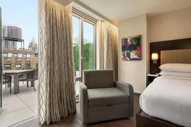 A hotel bedroom with a king bed, an armchair by the window, and a balcony with a table and chairs visible outside with city buildings beyond.