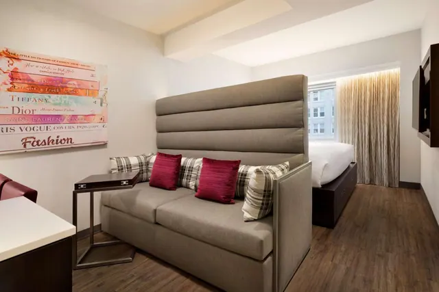 A hotel room with a tall grey upholstered sofa with red and plaid cushions, a bed beyond it, and a wall-mounted television.