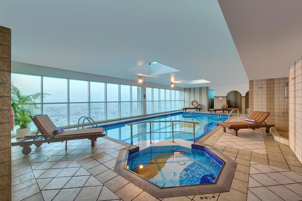 An indoor pool with floor-to-ceiling windows overlooking a hazy city view, with an octagonal hot tub and sun loungers along the edges.